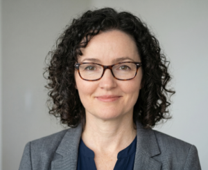 Portrait of a woman with curly dark hair wearing tortoiseshell glasses and a gray blazer against a neutral background, smiling subtly.