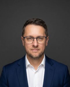 Professional head-and-shoulders portrait of a man in a navy suit and white shirt, wearing glasses, against a dark gray background.