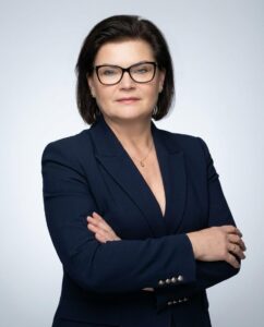Professional woman with short dark hair and glasses, wearing a navy blazer, arms crossed, looking at the camera.