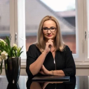 Professional-looking woman with glasses seated at a desk, hands resting near her chin, a laptop in front, flowers on the left, bright office window behind her.